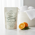 Roots & Leaves Powdered Laundry Detergent on white surface with laundry basket, white towels, rosemary and orange